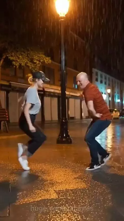 Couple dancing joyfully in the rain on a wet city street at night under a streetlight.