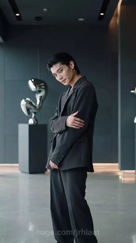 Young Asian man in a black suit jacket, black pants, and white sneakers posing confidently in a modern interior with silver sculptures.