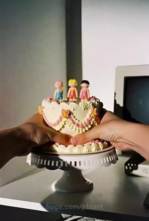 Close-up of a small white and pink cake with doll figures being squished by hands.