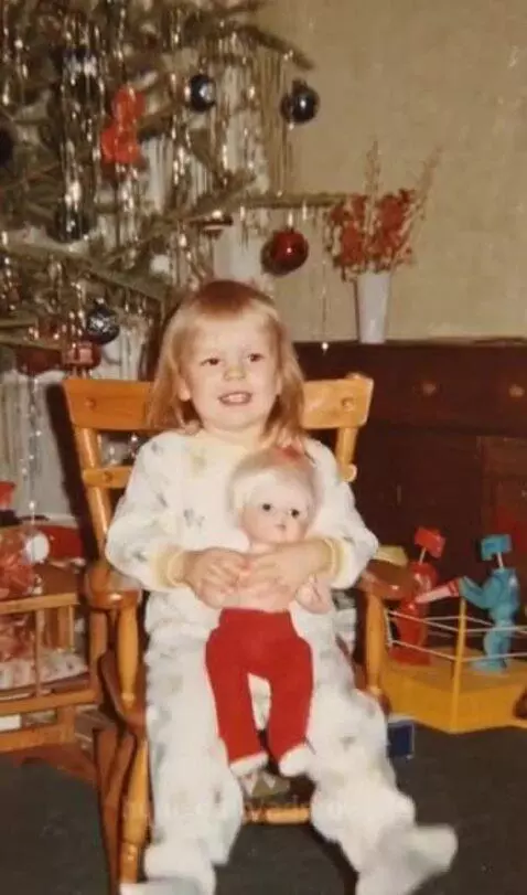 Young blonde girl in Christmas pajamas holding a doll while sitting in a rocking chair with a decorated Christmas tree.