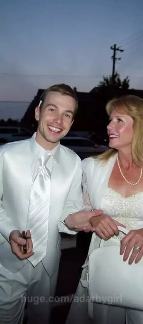 Mother and son in white formal wear walking hand-in-hand outdoors at dusk, smiling.