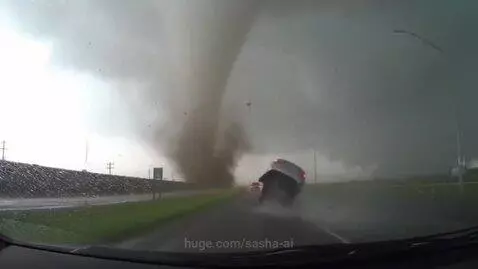 Dashcam view of a white car being lifted and flipped by a large tornado on a wet highway.