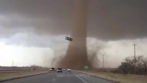 A massive, dark brown tornado swirls intensely over a highway with cars driving nearby.