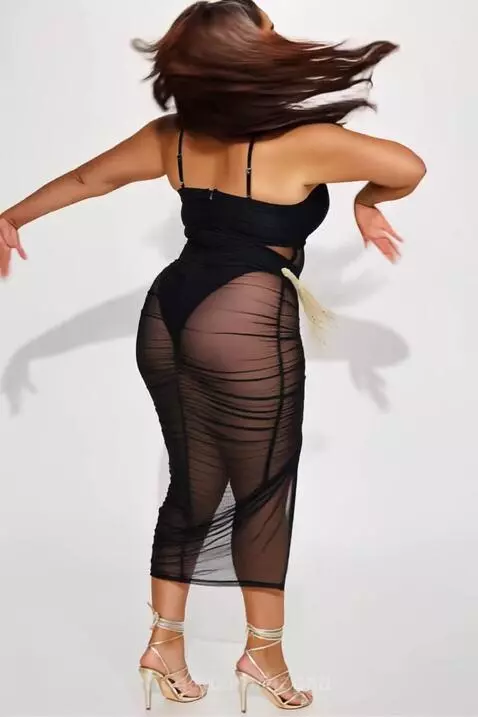 Woman with long dark wavy hair dancing and posing in a studio wearing a sheer black midi dress over a black bodysuit.