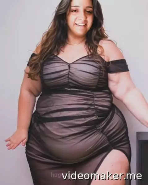 Plus-size woman with long dark hair smiling and posing in a sheer black off-the-shoulder dress with a high slit.