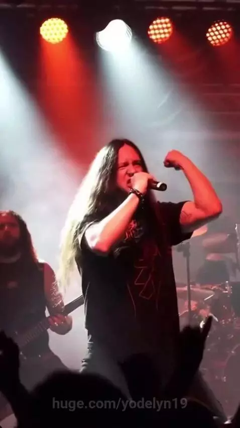 Death metal band performing live on a dimly lit stage with red and white spotlights, lead singer screaming into microphone.
