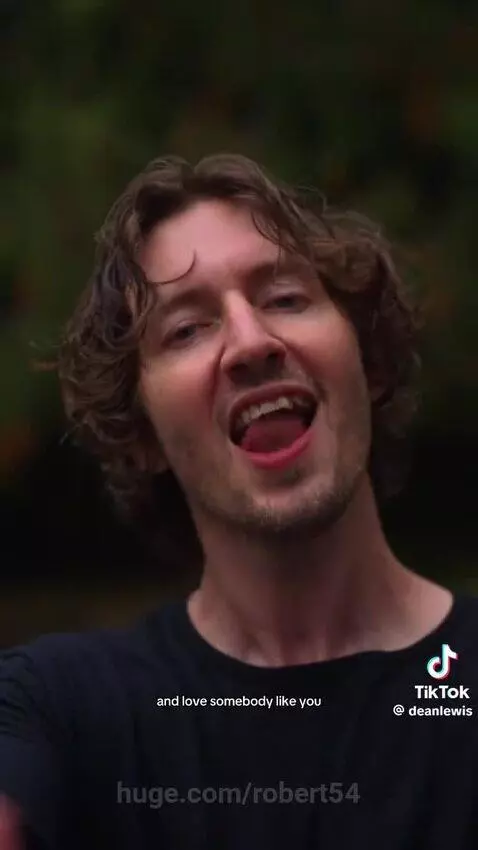 Man with curly brown hair and beard wearing a black t-shirt singing outdoors with a blurred green background.