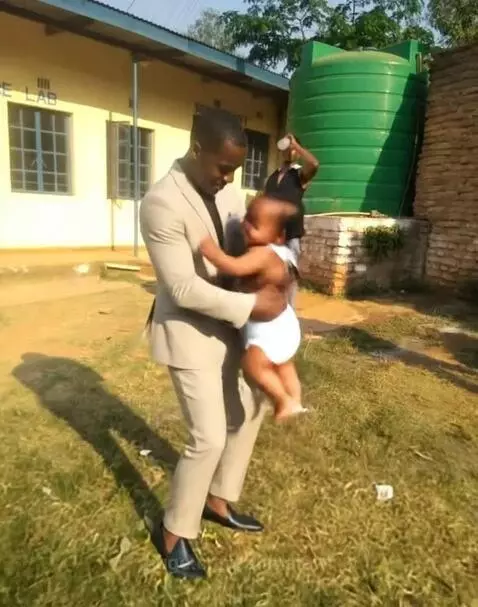 Man in a light suit holding a smiling baby, with a yellow 'SCIENCE LAB' building and green water tank in the background.