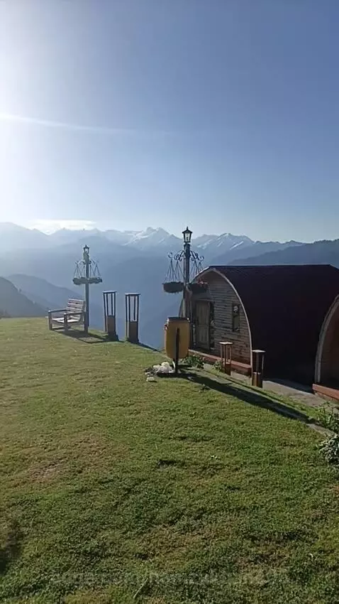 Rustic wooden cabins with red roofs on a green slope, with sunlit snow-capped mountains in the background under a blue sky.