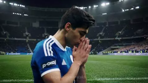 Young football player in blue and white jersey dribbling a football on a stadium pitch.