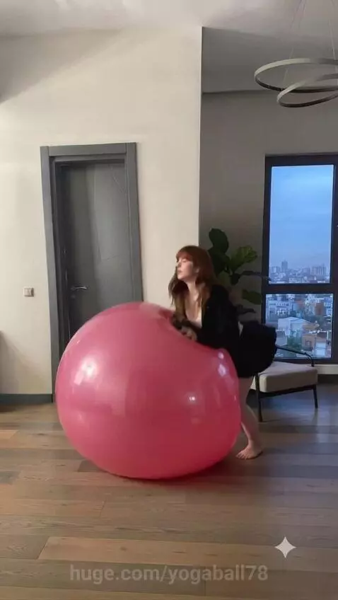 Woman in black and white outfit bouncing energetically on a large pink yoga ball in a modern apartment with city view.
