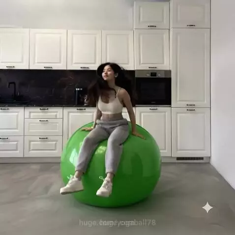 Woman with dark hair in a white sports bra and grey sweatpants bouncing on a large green yoga ball in a modern kitchen.