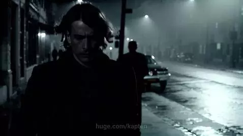 Man with long hair walks down a foggy, wet city street at night in black and white, illuminated by streetlights.