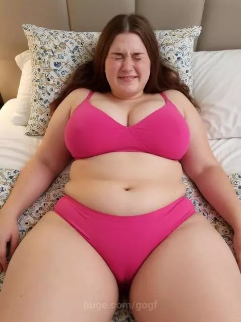 A full-figured woman with long brown hair lies on her back in bed, showing signs of intense labor pain with eyes squeezed shut and a contorted expression.