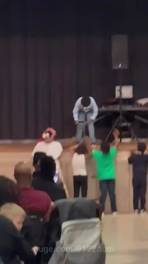 Performer in a blue shirt and headband on stage speaking into a microphone to an audience of children and adults.
