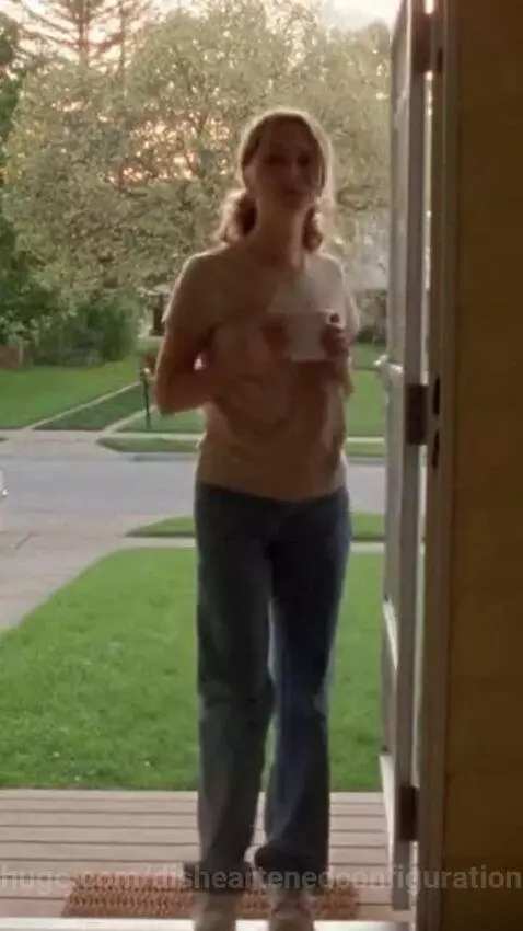 Young woman in a beige t-shirt and blue jeans steps onto a porch holding a coffee mug.