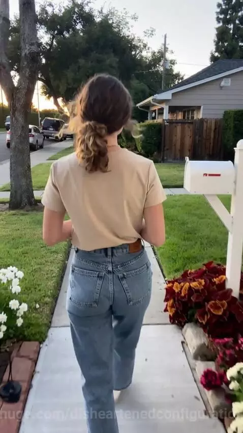 Young woman in a beige t-shirt and jeans walks down a suburban path with a coffee mug, seen from behind.