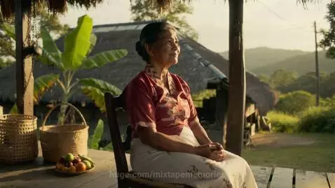 Elderly Filipino woman sitting peacefully on a wooden porch with woven baskets and fruits, rural landscape in background.