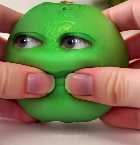 A green, round toy with human-like eyes and a smiling mouth being squeezed by hands.