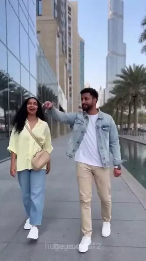 A man and woman walk in a modern city, pointing at a tall skyscraper with glass buildings and a water feature nearby.