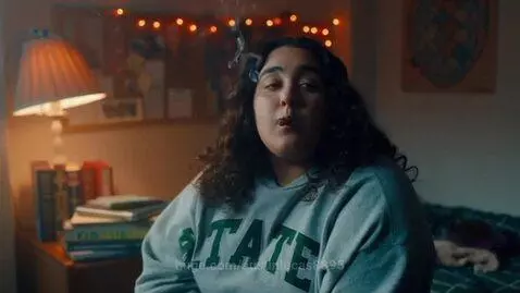 Young woman with curly dark hair wearing a grey sweatshirt, smoking a joint in a dimly lit room with string lights.