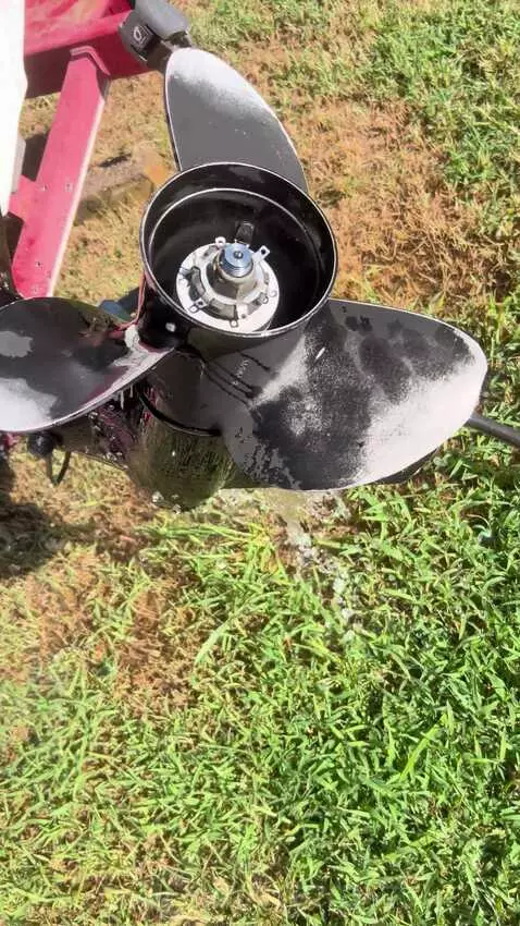 Close-up of a black outboard motor propeller being rinsed with water, with grass and a truck in the background.
