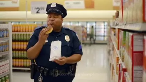 Police officer in uniform eating a donut in a supermarket aisle, smiling.