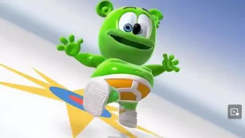 A smiling green gummy bear in underwear and shoes surfs on a target board, waving against a bright blue background.